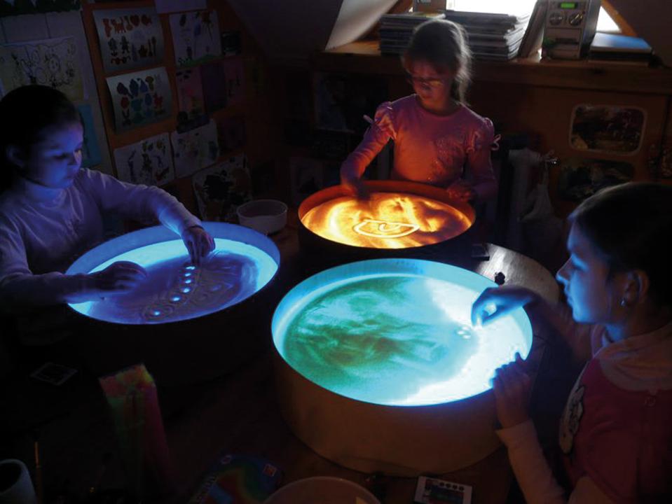 Leuchtkübel - drei Kinder malen mit Sand unterschiedliche Bilder in blau, gelb und grün. KiTa-Spielewelt