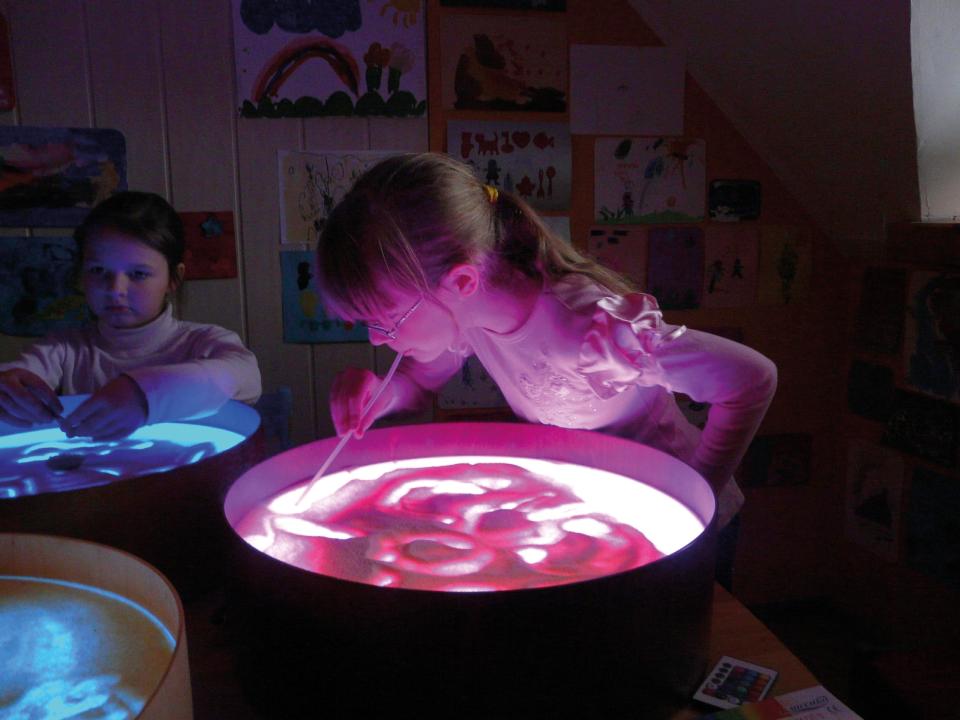 Leuchtkübel - zwei Kinder malen mit Sand und einem Pinsel auf der transparenten Oberfläche. KiTa-Spielewelt