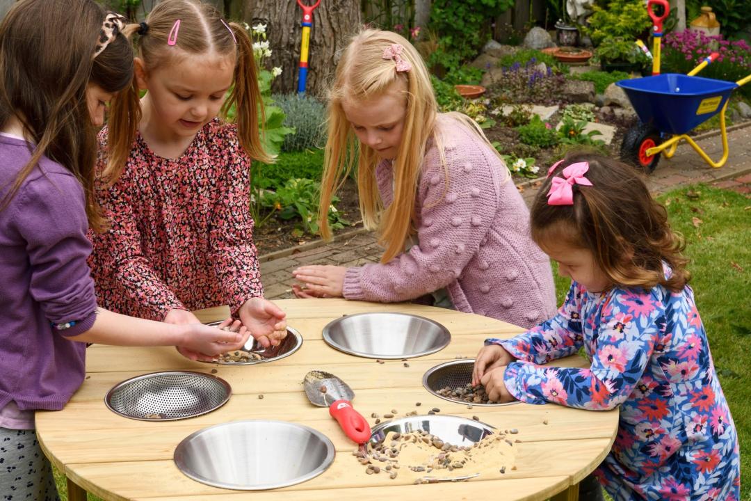 First Tools Outdoor Runder Sand-Spieltisch: Kinder spielen mit verschiedenen Sandelsachen am Holztisch. KiTa-Spielewelt