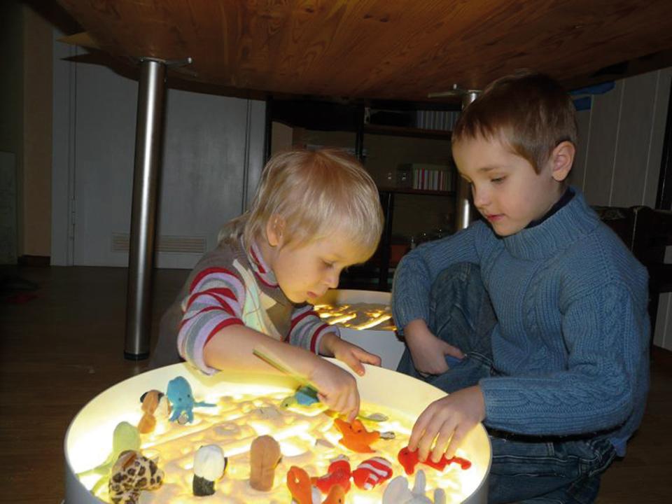 Leuchtkübel - zwei Kinder gestalten ein Bild mit Sand und Figuren, die von unten angestrahlt werden. KiTa-Spielewelt