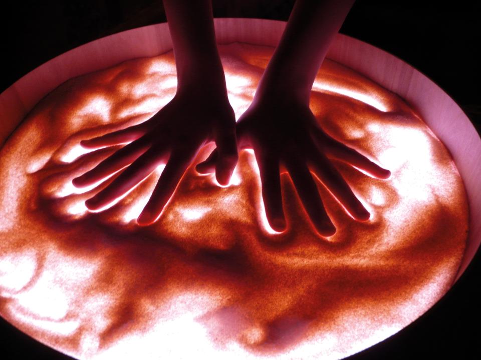 Leuchtkübel - Sandmalen mit den Händen auf transparenter Oberfläche, die von unten angestrahlt wird. KiTa-Spielewelt
