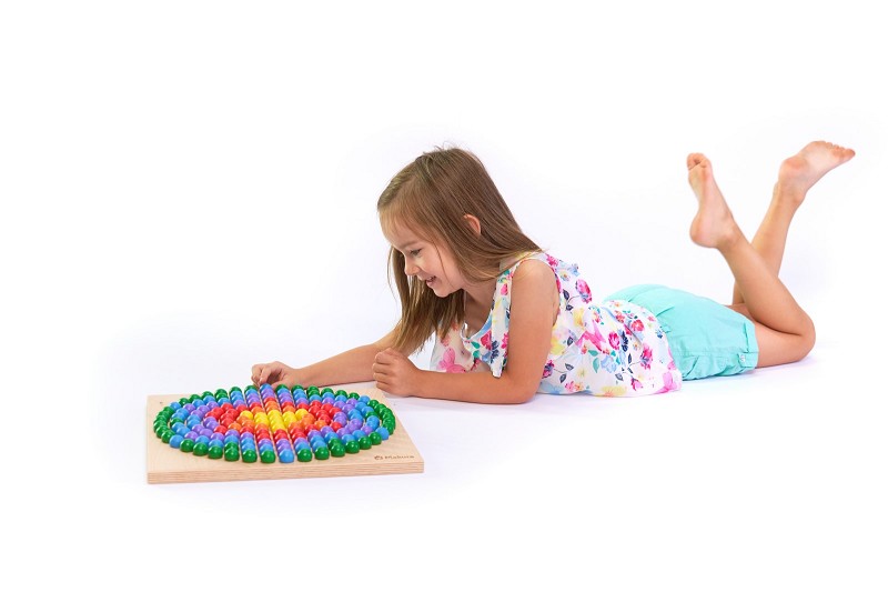 Kugelbrettspiel - Kugelbrettspiel: Ein Mädchen im Blumenshirt liegt auf dem Boden und spielt konzentriert mit einem bunten Makura Steckspiel, t
